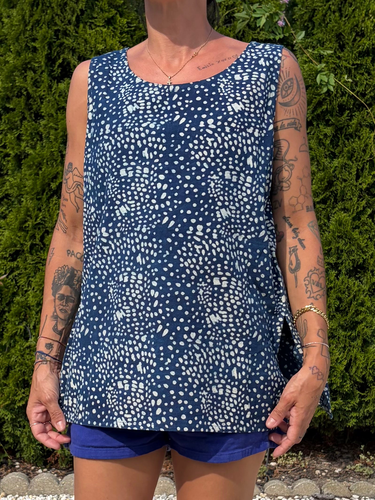 A woman wearing a sleeveless scoop-neck top with a navy blue and white speckled pattern, paired with blue shorts, standing outdoors in front of a green hedge.