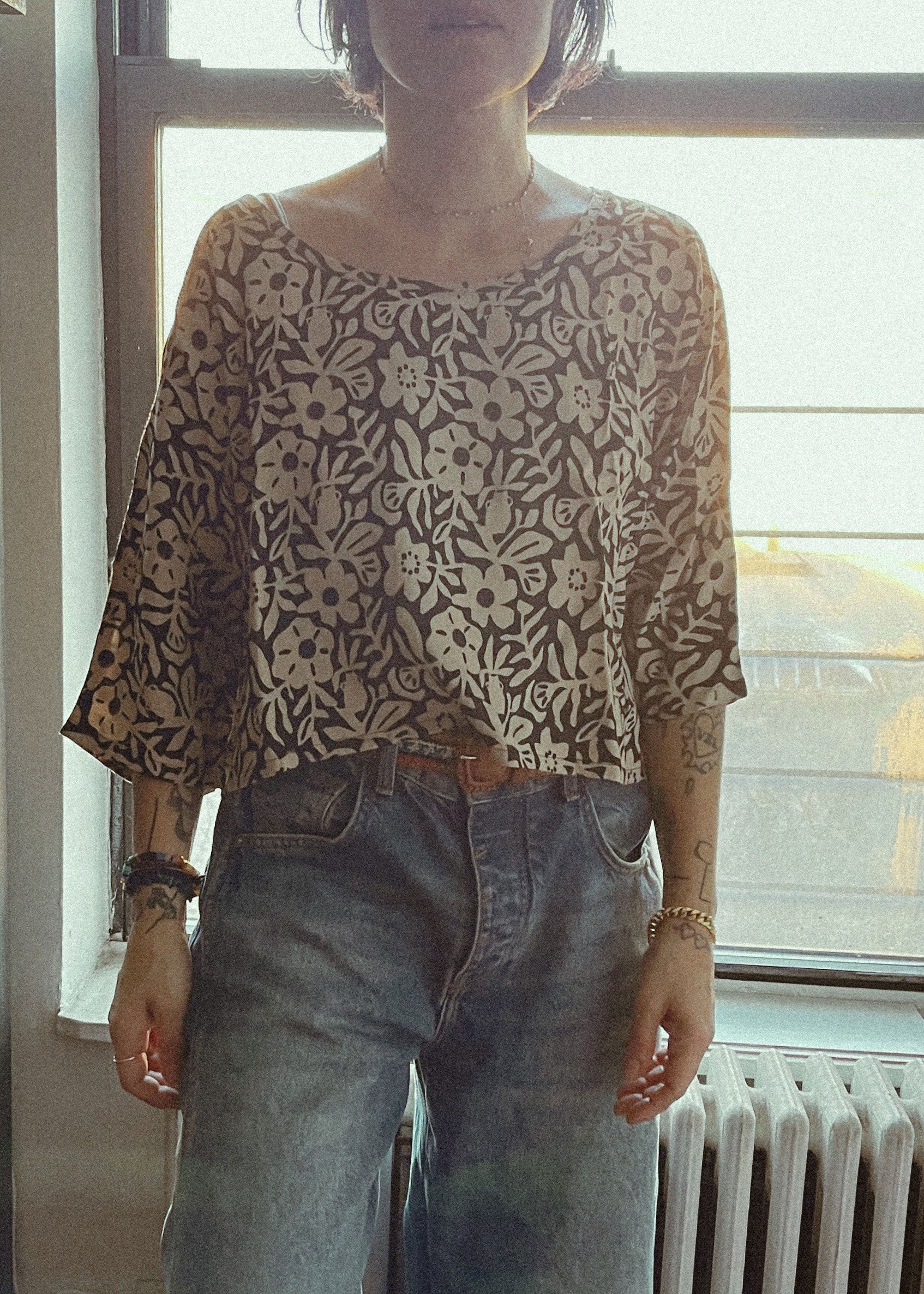 Person wearing a patterned top and jeans standing in front of a window.