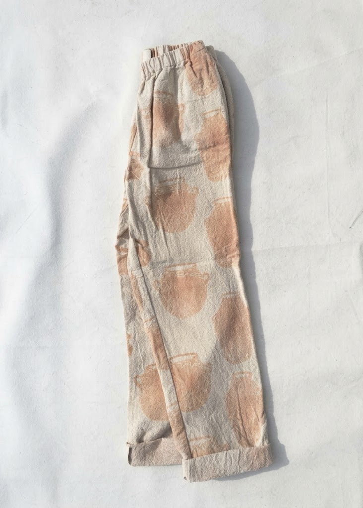 A pair of loose, cropped pants in a light beige linen-like fabric with a soft peach-pink abstract watercolor dot pattern, laid flat on a white background.