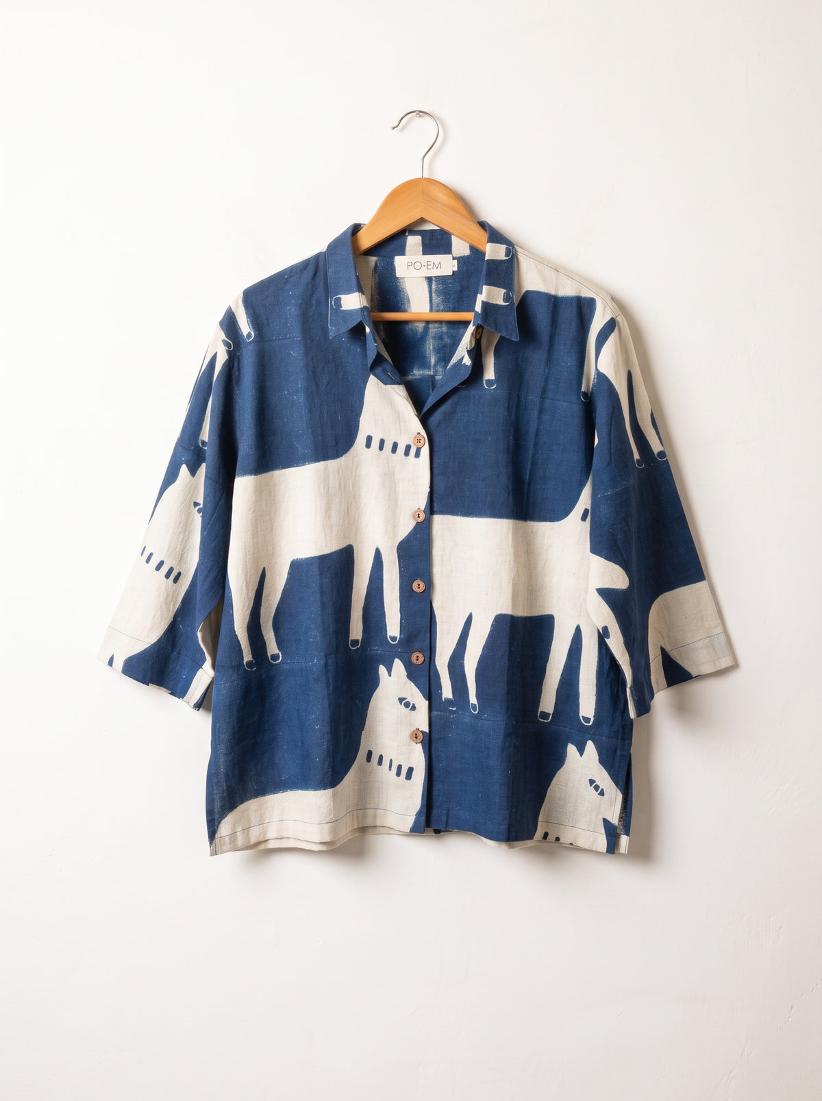 A long-sleeve button-up shirt with a blue and off-white horse print, hanging on a wooden hanger against a plain white wall by PO-EM slow fashion