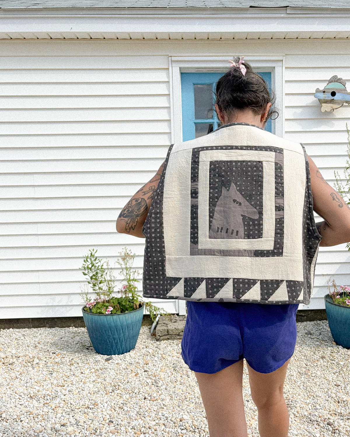A person viewed from behind wearing a sleeveless quilted vest outdoors in front of a white house. The vest has a large central square panel with a gray wolf head motif on a black and white patterned background, surrounded by cream and black geometric border sections.
